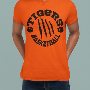 Puede incluir: Camiseta naranja con un gr&aacute;fico circular negro que dice "Tigers Basketball" con huellas de patas y marcas de garras.