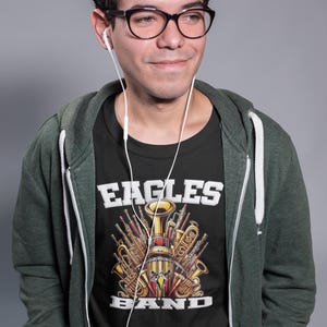 K&ouml;nnte beinhalten: Eine Person tr&auml;gt ein schwarzes T-Shirt mit einer Grafik von Musikinstrumenten und dem Text "Eagles Band". Die Person tr&auml;gt auch einen gr&uuml;nen Kapuzenpullover und hat wei&szlig;e Ohrh&ouml;rer in den Ohren.