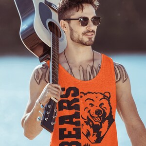 Pode incluir: Um homem usando &oacute;culos de sol e uma camiseta laranja sem mangas com um gr&aacute;fico de urso preto e a palavra "BEARS" nela. Ele est&aacute; segurando um viol&atilde;o marrom sobre o ombro.