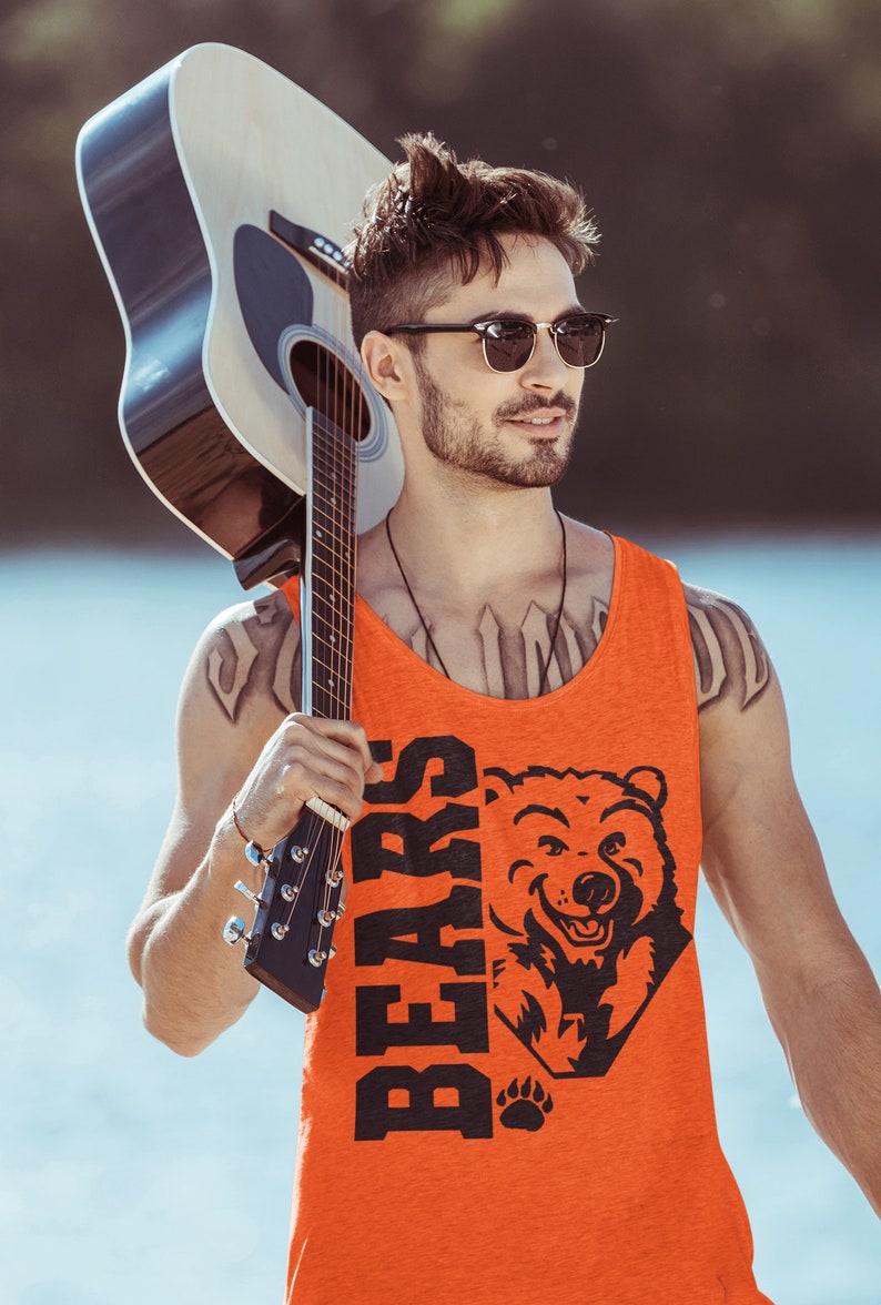 Pode incluir: Um homem usando &oacute;culos de sol e uma camiseta laranja sem mangas com um gr&aacute;fico de urso preto e a palavra "BEARS" nela. Ele est&aacute; segurando um viol&atilde;o marrom sobre o ombro.