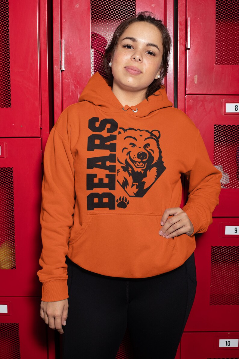 Pode incluir: Uma pessoa vestindo um moletom laranja com um gr&aacute;fico de urso preto e a palavra "BEARS" impressa na frente. O moletom est&aacute; na frente de um arm&aacute;rio vermelho.