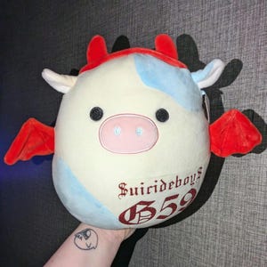 Peut inclure: Une peluche en forme de vache avec un corps blanc, des taches bleues et des détails rouges. Elle a des yeux noirs, un nez rose, des cornes et des ailes rouges. Le texte "Suicideboys 559" est imprimé en rouge.