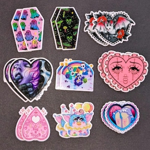 May include: Nine colorful stickers with various designs, including a heart with a crying face, a bat with a heart, a rainbow with ghosts, a heart with the text "Stay in Touch", a pink coffin with ghosts, a blue heart with a creature, a pink heart with a face, a blue heart with a creature, and a pink coffin with skulls.
