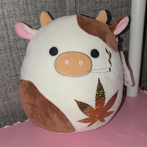May include: A brown and white plush cow with a brown marijuana leaf and a cigarette on its side. The cow has a pink nose and black eyes.