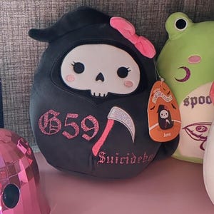 May include: Black plush toy shaped like a grim reaper with a pink bow, a skull face, and a pink scythe. The toy has pink glitter accents and the text "659" and "Suicideboy" on it. The toy is sitting on a pink surface next to a green frog plush toy.