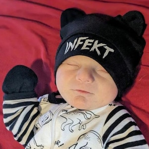 Op de afbeelding: Een baby draagt een zwarte muts met berenoren en het woord "INFEKT" in het wit. De baby draagt ook een zwart-wit gestreepte onesie met dinosaurusafbeeldingen.