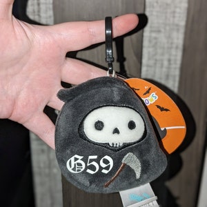 May include: A grey plush keychain shaped like the Grim Reaper with a white skull and a scythe. The keychain has the number "659" embroidered on the front.