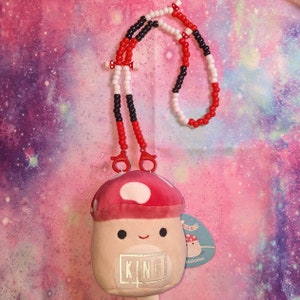May include: A mushroom-shaped plush toy with a red and white spotted cap, the word "KING" is embroidered on the front. It has a beaded lanyard in red, black, and white. The background is a colorful galaxy design.