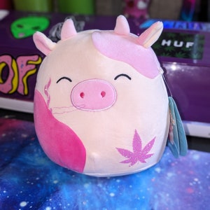 May include: Pink and white plush cow with a pink nose and a sparkly pink marijuana leaf on its side. The cow is smiling and has a tag attached to its ear.