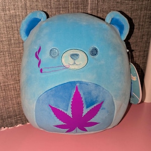 Może przedstawiać: Jasnoniebieski pluszowy miś z fioletowym liściem marihuany na brzuchu. Miś ma małą, haftowaną twarz i pali jointa. Pluszowa zabawka jest miękka i przytulna.