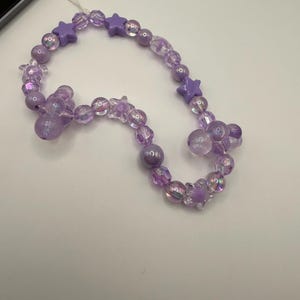 Può includere: Un ciondolo per telefono con perline con una varietà di perline viola, tra cui perline rotonde e a forma di stella. Le perline sono in tonalità lavanda e viola iridescente, infilate su un cordino trasparente.