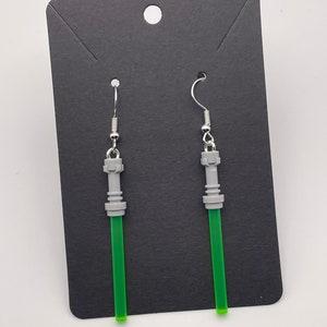 Puede incluir: Un par de pendientes de sable de luz de color plateado y verde. Los pendientes tienen forma de sable de luz con un mango plateado y una hoja verde.