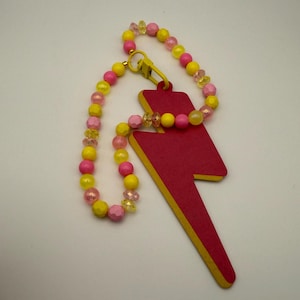 May include: A pink and yellow beaded keychain with a red lightning bolt charm. The keychain is made of round beads and has a gold metal clasp.