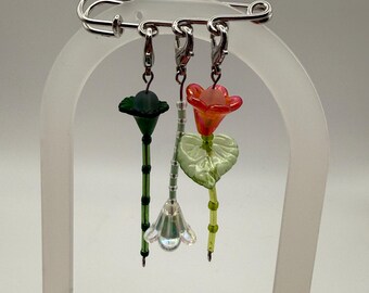 May include: A silver safety pin with three decorative floral charms. The charms feature a dark green flower, a clear flower, and a red and orange flower with a green leaf. The charms are made of glass and beads.