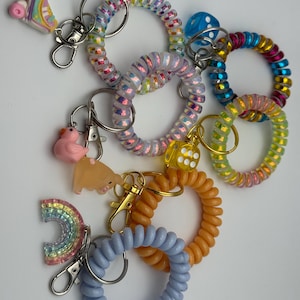 Puede incluir: Una colección de coloridos llaveros en espiral con varios dijes. Los llaveros presentan una variedad de colores, incluyendo azul, naranja, rosa e iridiscente. Los dijes incluyen un patín, un pato, un arcoíris y un dado.