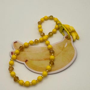 May include: A yellow beaded bracelet with a banana charm. The bracelet has round yellow beads and faceted amber-coloured beads. The charm is a yellow banana with a clasp. The bracelet rests on a banana-shaped card.