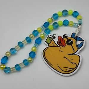 May include: A yellow rubber duck wearing sunglasses and a blue baseball cap, holding a green drink with a straw. The duck is attached to a beaded necklace with blue and yellow beads.
