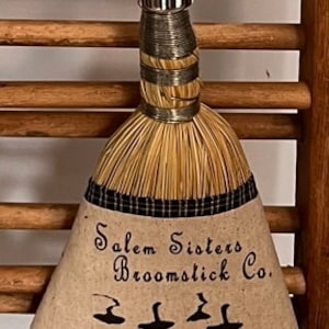 May include: A small, brown, straw broom with a black and white checkered ribbon hanging from the top. The broom has a white fabric label with the text "Salem Sisters Broomstick Co." and three black witch silhouettes.