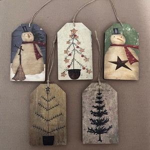 May include: Five Christmas-themed gift tags with various designs. Two tags feature a snowman with a black top hat and red scarf. One tag has a green background, and the other has a blue background. Three tags feature a stylized Christmas tree with a star on top. One tree is brown, one is green, and one is black.