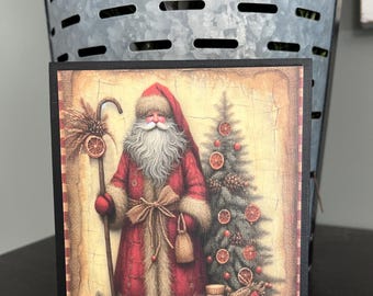 May include: A square Christmas-themed wooden block featuring a vintage illustration of Santa Claus in a red coat with fur trim, holding a staff. A Christmas tree and dried orange slices are also depicted.