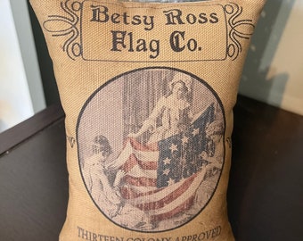 May include: A tan fabric pillow with a vintage image of a woman holding a large American flag. The pillow has the text "Betsy Ross Flag Co." and "Thirteen Colony Approved" printed on it.