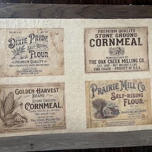 May include: A beige fabric featuring four vintage-style food labels. The labels include "Dixie Pride Self-Rising Flour," "Premium Quality Stone Ground Cornmeal," "Golden Harvest Brand Stone Ground Cornmeal," and "Prairie Mill Co. Self-Rising Flour."