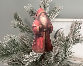 May include: A vintage-style Christmas ornament featuring a red-suited Santa Claus with a white beard and a white fur trim. The ornament is hanging on a snowy artificial Christmas tree.