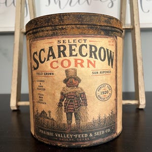May include: A cylindrical metal tin with a distressed, rusted lid and a tan label featuring the text "Select Scarecrow Corn." The label displays a graphic of a scarecrow in a field, "Prairie Valley Feed & Seed Co. Dallas, Texas," and "Net Wt 56 Lbs."