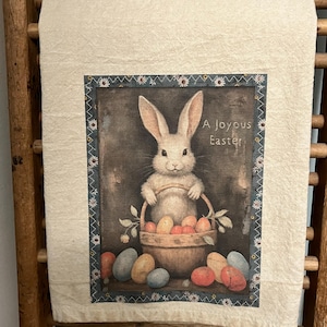 May include: A white bunny rabbit holds a basket of colorful Easter eggs. The bunny is sitting on a brown surface with more eggs scattered around the basket. The image has a vintage feel and includes the text "A Joyous Easter".
