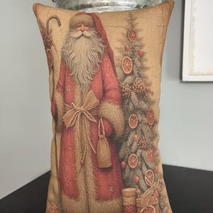 May include: Rectangular decorative pillow featuring a vintage illustration of Santa Claus in a red coat, holding a staff and bag. A Christmas tree with orange slices is to the right. The pillow has a beige background.