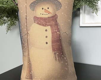 Pode incluir: Uma almofada bege com uma ilustração de boneco de neve e o texto "Winter Welcome". O boneco de neve está usando uma echarpe vermelha e preta e um chapéu-coco preto. O fundo é marrom claro com flocos de neve brancos.