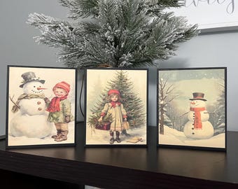 May include: Three framed vintage Christmas illustrations. The first shows a snowman with a child holding a red scarf. The second shows a girl in a red hat standing next to a Christmas tree. The third shows a snowman in a winter scene.