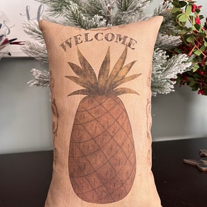 May include: A tan pillow with a brown pineapple and the word "WELCOME" printed on it.