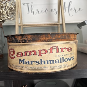 May include: A vintage metal container of Campfire Marshmallows. The label features the words "Campfire Marshmallows" in red and blue, with "The Original Food" above. The container has a rusty, aged appearance.