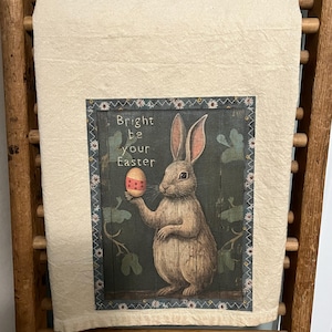 May include: A beige tea towel with a printed image of a rabbit holding a decorated Easter egg. The image is surrounded by a border of white flowers and green leaves. The text "Bright be your Easter" is printed above the rabbit.