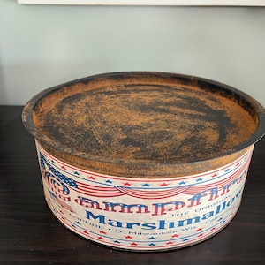 May include: Vintage Campfire Marshmallows tin with a distressed, rusty lid. The tin features a red, white, and blue design with the American flag and text that reads "Campfire Marshmallows, The Original". The tin is cylindrical and sits on a dark surface.
