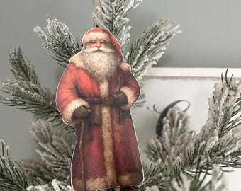 May include: A vintage-style Santa Claus ornament with a red and white suit, white beard, and a staff. The ornament is hanging on a snowy artificial Christmas tree.