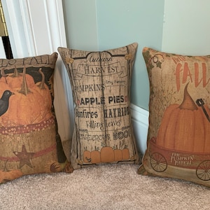 May include: Three decorative throw pillows with fall-themed designs. The pillows are made of a burlap-like fabric and feature images of pumpkins, birds, and fall-themed text. The text on the pillows reads "Hello Fall", "Autumn Harvest", and "Hoo Fall".