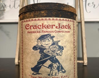 Può includere: Latta vintage di Cracker Jack con finitura invecchiata. L'etichetta presenta un ragazzo marinaio con un cane e il testo "Cracker Jack" e "Americas Famous Confection". La latta presenta anche il testo "The More You Eat - The More You Want".