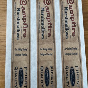 May include: Three vintage-style labels for Campfire Marshmallows, featuring a brown and cream color scheme. The labels include the text "Campfire Marshmallows," "The Original Food," and "Finest Quality." They are designed for baking, topping, eating, and toasting.