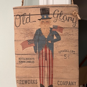 May include: A tan burlap sign with a wood grain pattern featuring a cartoon Uncle Sam holding two American flags. The sign reads "Old Glory Fireworks Company" and "Sparklers 5¢".  The sign also includes the text "Bottle Rockets Roman Candles".