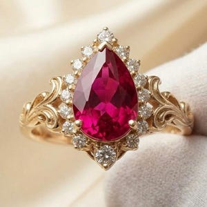 Pode incluir: Um anel de ouro com uma grande pedra preciosa vermelho-escura em forma de pera no centro, rodeada por um halo de pequenos diamantes brancos redondos. A banda apresenta detalhes de volutas em ambos os lados, em metal ouro amarelo polido.