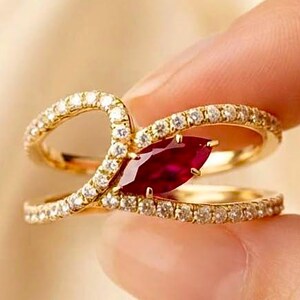 May include: A gold ring featuring a marquise-cut deep red gemstone at the center, flanked by two bypass-style bands encrusted with small clear stones. The design creates an open, overlapping silhouette with a polished metallic finish throughout.