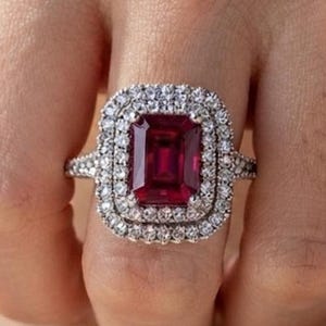 Peut inclure: Une bague ornée d'une grande pierre précieuse rectangulaire rouge foncé, entourée d'un double halo de petites pierres rondes transparentes. L'anneau est métallique et incrusté de minuscules pierres claires sur les côtés, offrant un éclat.