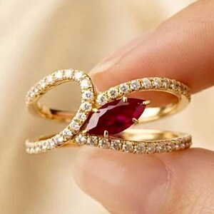 Peut inclure: Une bague en or ornée d'une pierre précieuse rouge foncé taille marquise au centre, entourée de deux anneaux incurvés incrustés de petites pierres claires. Le design crée une silhouette ouverte et superposée avec une finition polie.