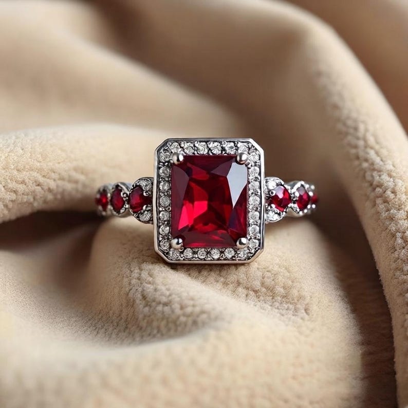 K&ouml;nnte beinhalten: Ein silberfarbener Ring mit einem gro&szlig;en, rechteckigen, facettierten roten Mittelstein, der von einem Kranz aus kleinen klaren Steinen umgeben ist. Die Ringschiene ist an den Seiten mit weiteren kleinen roten und klaren Steinen verziert.