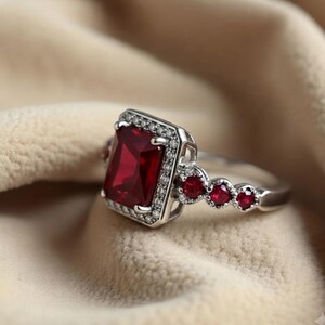 K&ouml;nnte beinhalten: Ein silberfarbener Ring mit einem gro&szlig;en, rechteckigen, tiefroten Mittelstein, der von einem Kranz aus kleinen, klaren, funkelnden Steinen umgeben ist. Die Ringschiene ist mit drei weiteren runden, tiefroten Steinen an jeder Seite verziert.