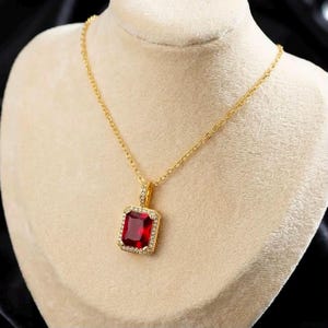 Colar elegante com pingente de pedra preciosa vermelha lapidada em esmeralda e halo de zircônias cúbicas em tom dourado.