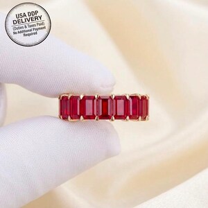 Könnte beinhalten: Ein Goldring mit einer durchgehenden Reihe von tiefroten Rubinen im Smaragdschliff, die in einer Krappenfassung gehalten werden. Der Ring wird zwischen zwei Fingern in weißen Stoffhandschuhen gehalten und zeigt die leuchtenden Edelsteine.