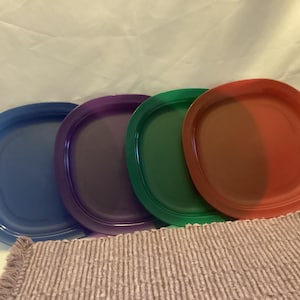 Könnte beinhalten: Vier farbige, ovale Teller in Blau, Lila, Grün und Rot. Die Teller sind in einer Reihe angeordnet, mit einem strukturierten, hellbraunen Platzset im Vordergrund. Die Teller bestehen aus glänzendem Material.